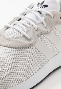 Valkoinen urheilukenkä, jossa verkkokangasta ja mokkanahkaisia yksityiskohtia. Kolme vaakasuoraa brändiviivaa ja valkoiset nauhat. Kaareva muotoilu.