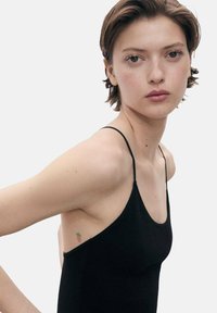 Black tank top with thin straps, featuring a fitted design and textured fabric. Small green strawberry tattoo visible on the left armpit.