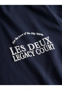 Tamsiai mėlynas medvilnės audinys su baltu siuvinėtų tekstu: "LES DEUX LEGACY COURT" ir "Dėl meilės Didžiajam Obuoliui," su tekstūruotu siuvimu.