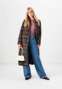 Blond kvinna står klädd i en brun rutigt kappa, en mauveblus, vida blå jeans, bordeauxskor och håller en liten vit handväska.