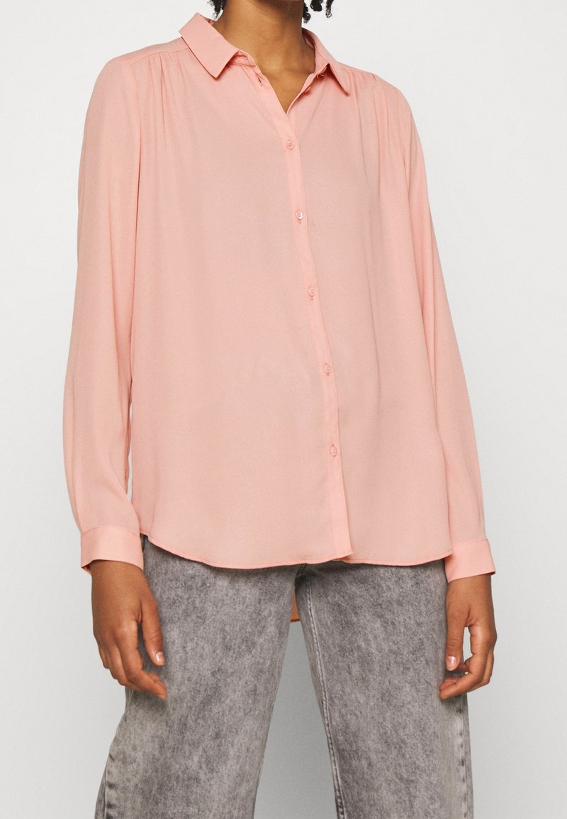 Personne portant une chemise ample pêche à manches longues boutonnée, associée à un jean gris délavé taille haute sur un fond uni.