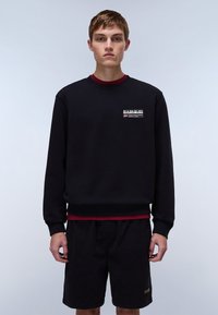 Sort sweatshirt med rød detalje ved halsudskæringen, der har en logo-patch foran. Båret med sorte shorts, sat mod en neutral baggrund.