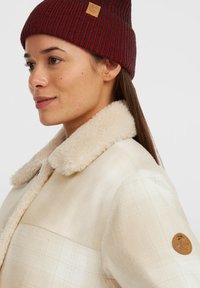 Kvinde iført et cremelangtternet frakke med et shearling krave og en ribbet mørkerød strikket hue, vendt til venstre med et svagt smil.