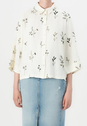 Blouse blanche boutonnée avec manches amples ornée d'une délicate broderie florale noire et grise, associée à une jupe en denim bleu clair avec une petite déchirure.