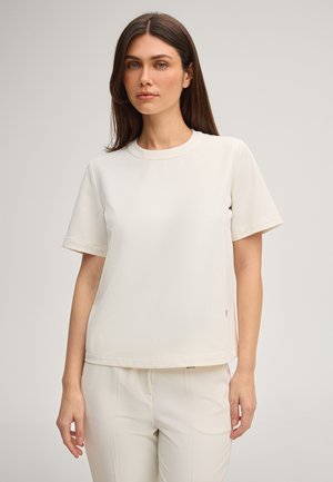 Młoda kobieta o długich ciemnych włosach, ubrana w prostą białą koszulkę z krótkim rękawem oraz białe spodnie, stojąca na neutralnym tle.