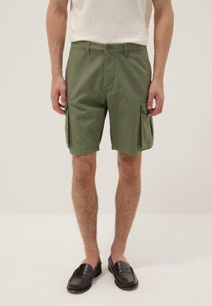 Hombre que lleva pantalones cortos cargo verde oliva con bolsillos laterales, camiseta blanca y mocasines negros, de pie frente a un fondo claro y liso.