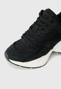 Schwarzer Sportschuh aus Mesh und Wildleder mit strukturierten Akzenten und einer klobigen weißen Sohle mit schwarzem Fußteil. Schnürdesign.