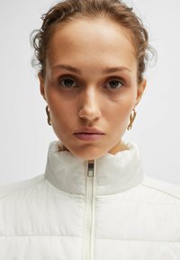Giacca imbottita bianca con colletto alto, dotata di chiusura frontale a zip e texture trapuntata. Orecchini a cerchio dorati completano il look.