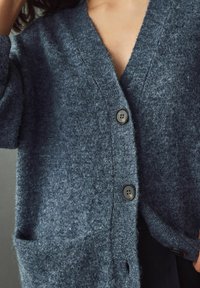 Mörkblå stickad cardigan med V-ringning, två framfickor och två svarta knappar. Tyget har en mjuk, texturerad yta.