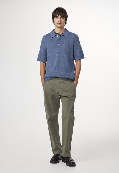 Polo a maniche corte, di colore azzurro chiaro, con colletto e tessuto texturizzato, abbinato a pantaloni verdi oliva e scarpe nere.
