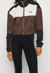 Kraftigt skuren jacka i brunt leopardtryck med vita och svarta paneler, metallisk dragkedja, stållkrage och elastiska ärmar. Matchad med svarta leggings.