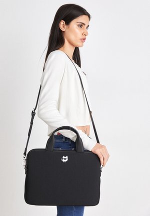 Nainen valkoisessa neuleessa ja farkuissa kantaa mustaa kannettavan tietokoneen laukku, jossa on kahvat ja olkahihna sekä pieni kissalogo.