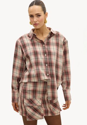 Femme portant une chemise oversize à carreaux rose et beige boutonnée, avec une jupe assortie à carreaux dotée d'une grande poche frontale, et des boucles d'oreilles anneaux dorés.