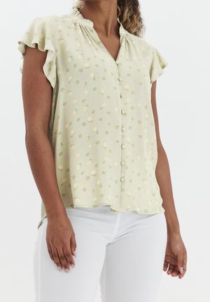 Femme portant un chemisier à manches courtes vert clair avec un petit imprimé floral et des boutons recouverts de tissu, assorti à un pantalon blanc.