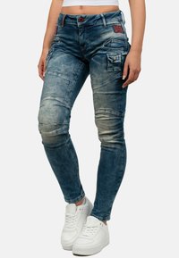 Jeans in denim azzurro chiaro con una vestibilità slim, caratterizzati da cuciture a pannello, molteplici tasche e una texture consumata. Indossati con sneakers bianche.