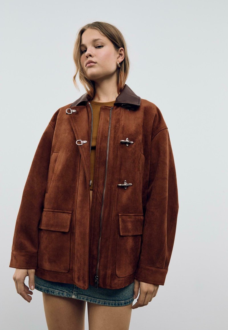 Veste en daim marron avec un col en cuir marron foncé, dotée de ferrures en métal argenté et de deux grandes poches avant ; design décontracté.