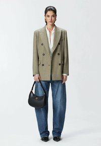 Blazer doppiopetto verde oliva con grandi bottoni neri, camicia bianca, jeans blu oversize, tiene in mano una borsa nera con dettagli dorati.