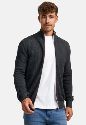 Cárdigan de zip oscuro con un patrón texturizado, cuello y puños acanalados, y detalles de cremallera en contraste, llevado sobre una camiseta blanca.