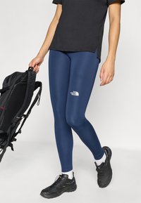 Στενά κολάν σε ναυτικό μπλε από ελαστικό υλικό, με το λευκό λογότυπο της The North Face στον αριστερό μηρό, συνδυασμένα με μαύρο σακίδιο πλάτης.