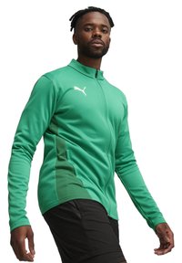 Groene sportjack met volledige rits, lange mouwen, zijpanelen van mesh en een witte logo-accent. Gecombineerd met zwarte shorts.