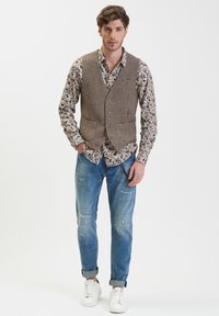 Gilet a quadri marroni sopra una camicia con motivo floreale, abbinato a jeans chiari denim strappati e sneakers bianche. Look semplice e strutturato.