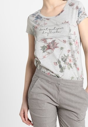 Femme portant un t-shirt gris floral avec le texte "don't quit your daydream" et un pantalon à carreaux, debout sur un fond clair uni.