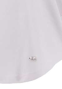 Witte T-shirt met een gladde textuur, een geschulpte onderrand en een klein zilveren metalen label met de tekst "KEY LARGO" in de linkerbenedenhoek.