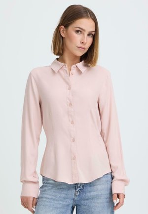 Mujer joven con cabello lacio que lleva una camisa de botones rosa pálido y jeans azul claro, posando frente a un fondo liso.