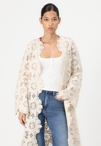Lace duster in cream, featuring floral patterns and scalloped edges. Pairing with a white tank top and blue jeans. Light, delicate texture.