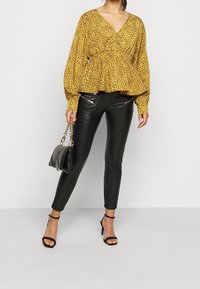 Gul leopardmönstrad blus med peplumsilhuett, i kombination med svarta skinnybyxor i konstläder och svarta remsandaler, som håller en svart väska.