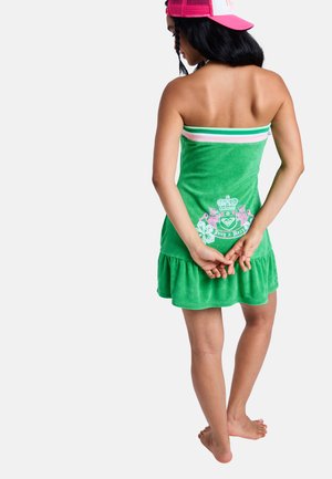 Frau trägt ein grünes trägerloses Kleid mit einem floralen Wappen, rosa und weißen Streifen oben und einer rosa Baseballkappe, steht barfuß.
