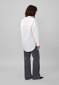 White, long-sleeve shirt with a pointed collar and curved hem, paired with loose, dark gray denim jeans. Smooth fabric texture is visible.