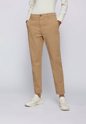 Personne portant un pantalon beige droit, des baskets blanches et un haut blanc à manches longues, les mains dans les poches, debout sur un fond uni.