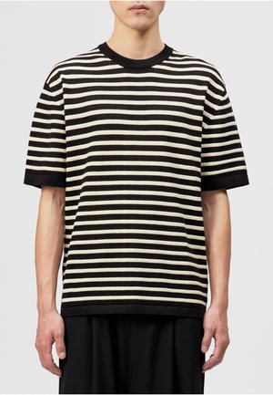 Mann trägt ein schwarz-weiß gestreiftes Kurzarmshirt mit rundem Ausschnitt, steht vor einem schlichten Hintergrund.