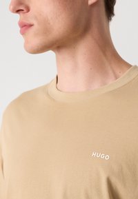 Bežs kokvilnas t-krekls ar apaļu kakla izgriezumu un mazu baltu "HUGO" logo kreisajā pusē. Gluda tekstūra, ikdienišķs piegriezums.