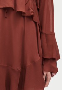 Robe en tissu transparent de couleur rouille avec des détails à volants superposés sur les manches et l'ourlet, portée sur le bras et la main d'une personne.