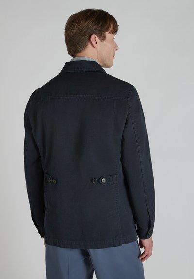 Homme portant une veste bleu marine foncé avec des sangles arrière boutonnées, de côté, associé à un pantalon bleu clair, sur un fond uni.