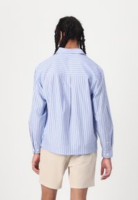 Camicia a righe blu e bianche con collare a bottoni, maniche lunghe e polsini a bottone; tessuto leggero, vestibilità leggermente rilassata.