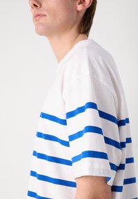 Weißes T-Shirt mit kräftigen blauen horizontalen Streifen. Kurzärmlig und mit lockerer Passform. Glattes Baumwollmaterial mit klassischem Rundhalsausschnitt.