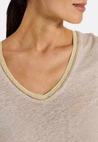 Beige V-Ausschnitt-Oberteil mit einem goldakzentuierten Halsauschnitt. Weicher, leichter Stoff mit einer subtilen Textur und lockerer Passform.
