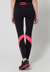 Persona che indossa leggings neri con accentuazioni curve rosa sui polpacci e una maglietta nera con una piccola tasca con zip rosa nella parte posteriore.