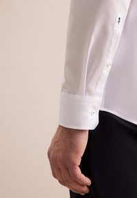 Camicia bianca con una trama strutturata, dotata di polsino con bottoni e dettaglio floreale ricamato sulla manica. Finitura in tessuto liscia.