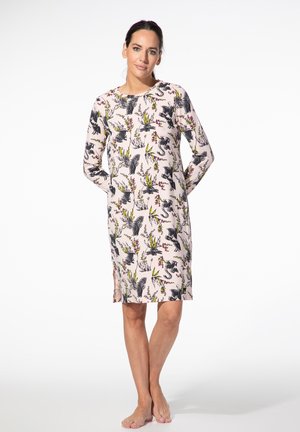 Woman standing barefoot wearing a long-sleeve light pink dress with black and yellow bird and plant prints.