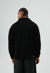 Zwarte fleecejas met een gestructureerd, pluizig materiaal, een stevige kraag en een licht oversized pasvorm met zichtbare stiksel details.