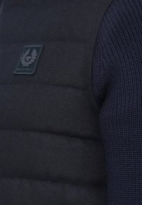 Detailný pohľad na čiernu vankúšikovu vestu s nášivkou loga Belstaff, oblečenou cez tmavomodrý pletený sveter.