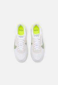 Zapatillas blancas con acentos verdes, parte superior de ante y cuero texturizado, puntera perforada, cordones blancos y forro interior amarillo brillante. Logotipo de Nike presente.