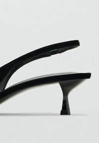 Svarta mocka klacksandaler med en smal remdesign, som har en glänsande svart klack och en minimalistisk form. Texturerat tyg framhäver stilen.