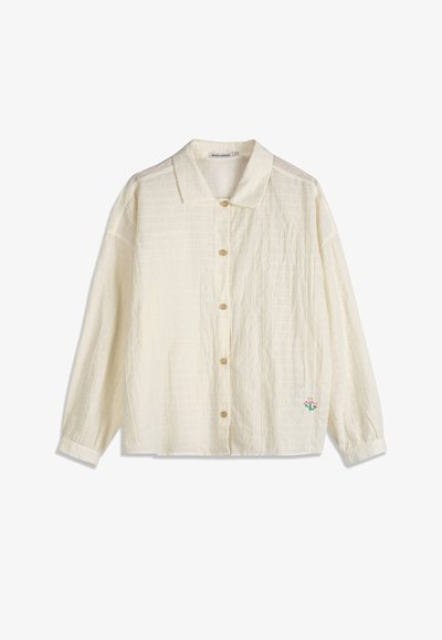 Chemise à manches longues couleur crème avec une texture subtile en grille et une petite fleur brodée rose et verte près de l'ourlet.