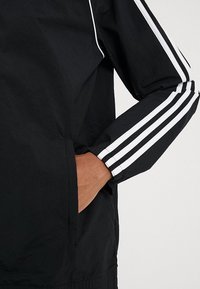 Černá bunda s bílými pruhy podél rukávů, s předním zipem a bočními kapsami. Materiál má hladkou, lehkou texturu.