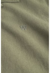 Polo shirt vert olive en tissu doux, avec un subtil logo 'W' brodé en gris clair. Texture lisse avec des détails de couture minimaux.